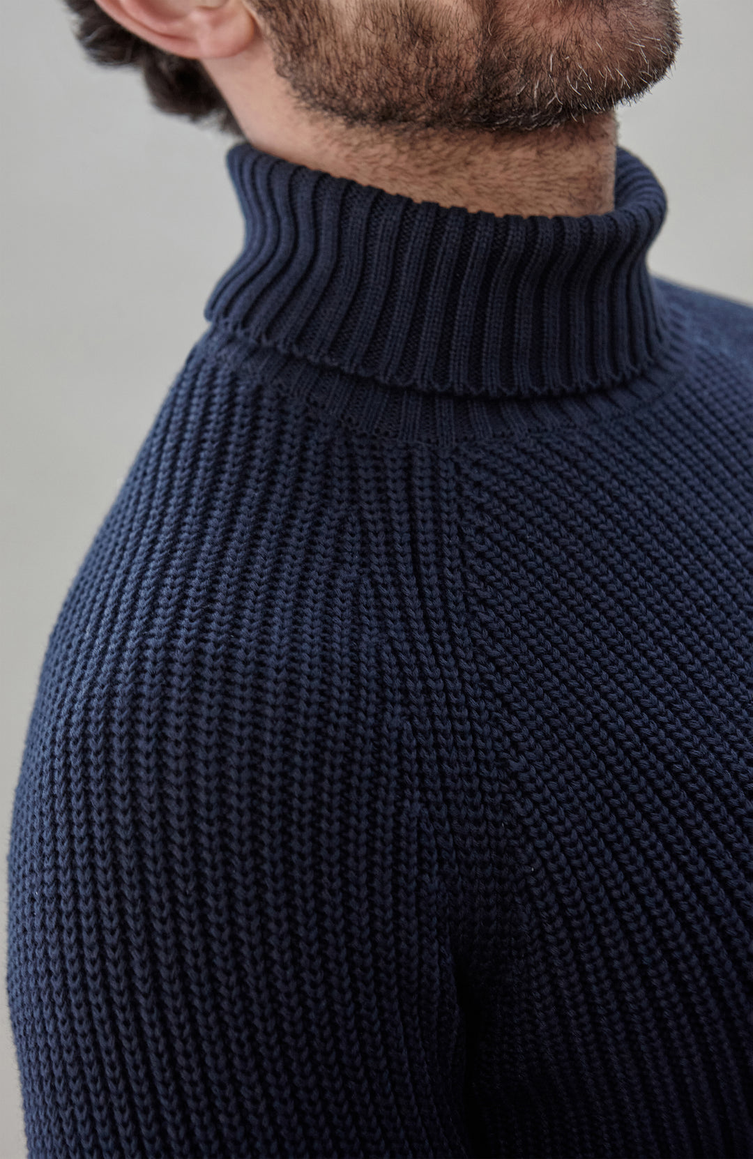 Man wearing a dark blue ribbed cotton roll neck sweater by Paul James Knitwear and brown pants holding an umbrella against a plain background