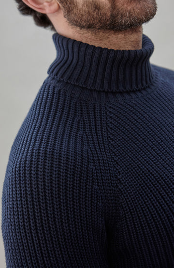 Man wearing a dark blue ribbed cotton roll neck sweater by Paul James Knitwear and brown pants holding an umbrella against a plain background