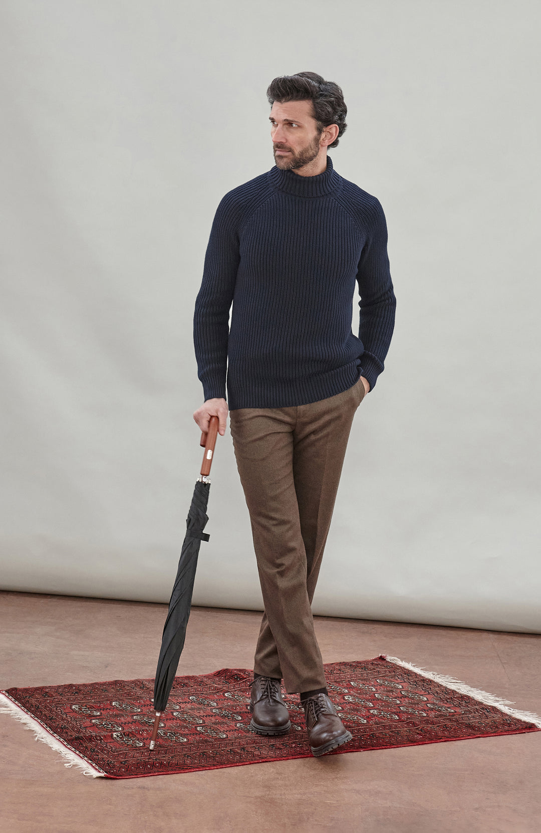 Man wearing a dark blue ribbed cotton roll neck sweater by Paul James Knitwear and brown pants holding an umbrella against a plain background