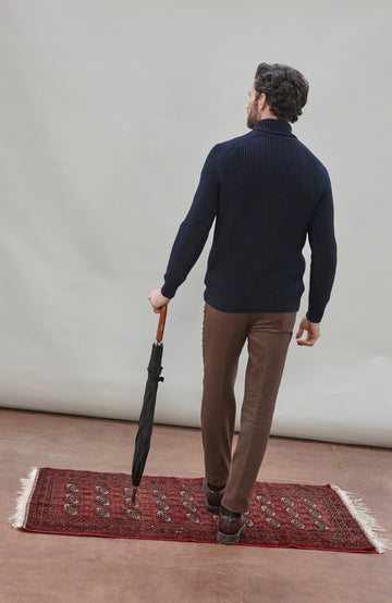 Man wearing a dark blue ribbed cotton roll neck sweater by Paul James Knitwear and brown pants holding an umbrella against a plain background