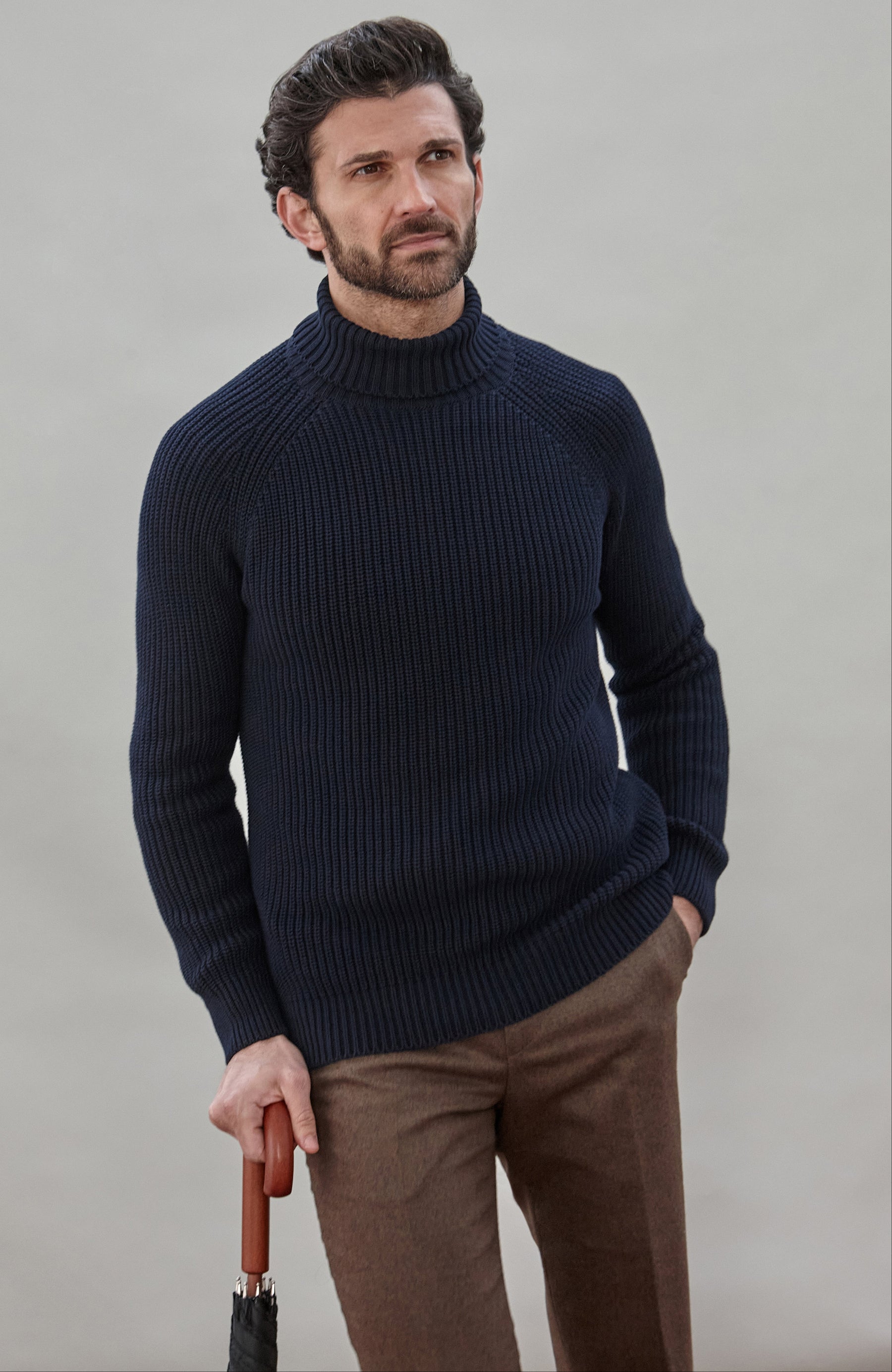 Man wearing a dark blue ribbed cotton roll neck sweater by Paul James Knitwear and brown pants holding an umbrella against a plain background