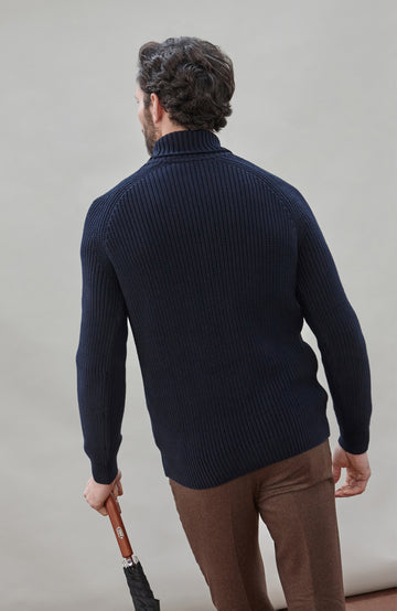 Man wearing a dark blue ribbed cotton roll neck sweater by Paul James Knitwear and brown pants holding an umbrella against a plain background
