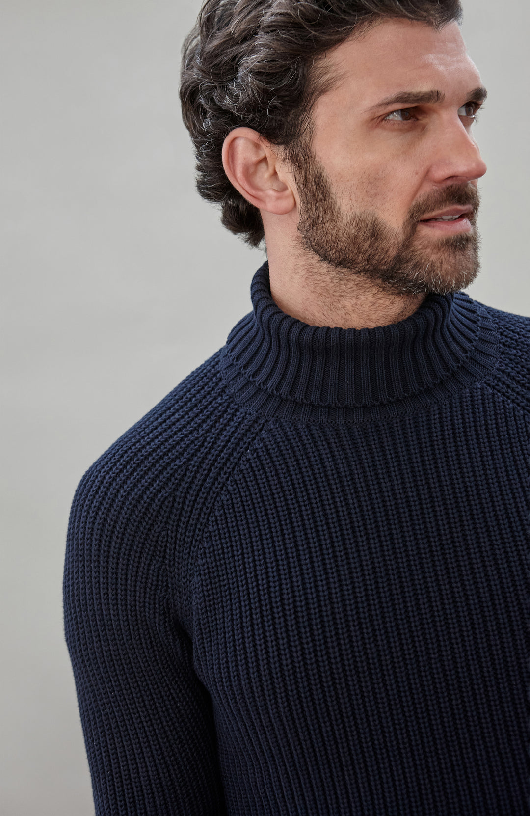 Man wearing a dark blue ribbed cotton roll neck sweater by Paul James Knitwear and brown pants holding an umbrella against a plain background
