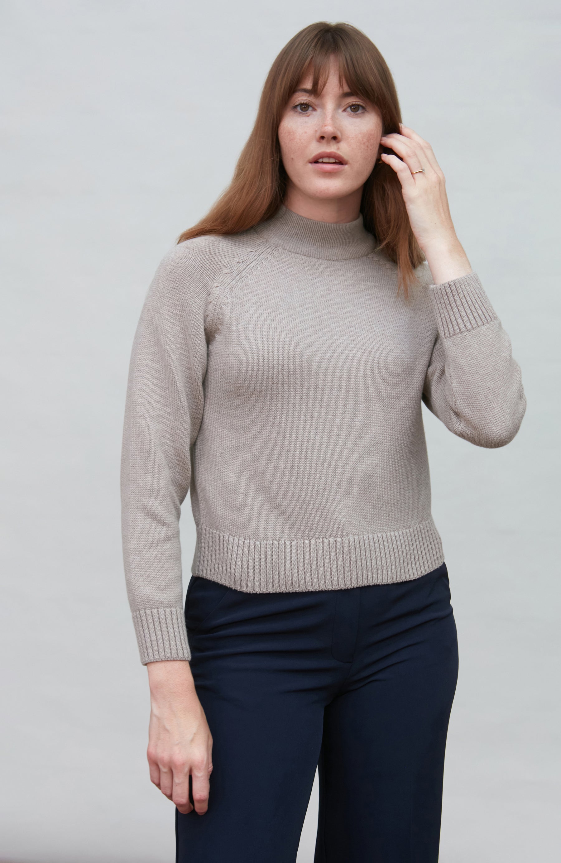Woman wearing a beige cotton mock neck paul james sweater and navy pants against a plain background