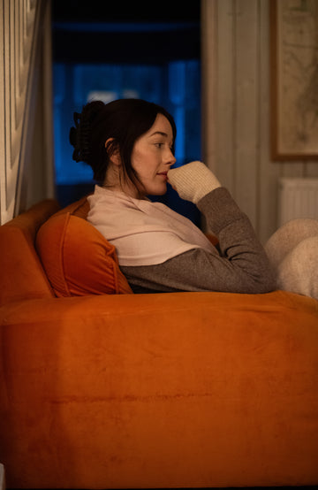 A woman in a cozy living room setting, reclining on an orange armchair and wearing a llight cream merino cashmere shawl, with a white knit glove covering part of her face.
