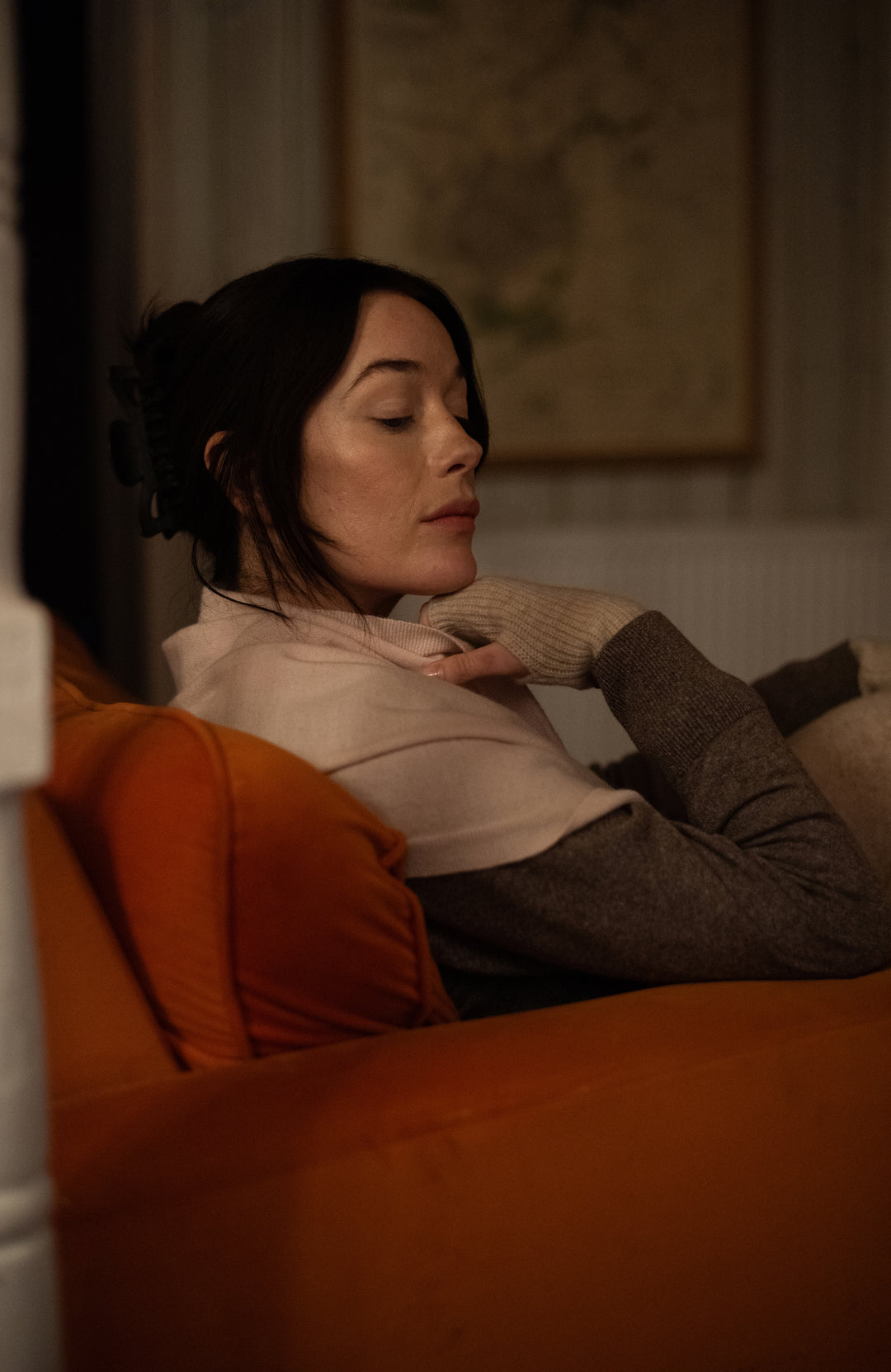 A woman reclining on an orange couch in a dimly lit room, wearing light cream merino cashmere shawl by Paul James Knitwear