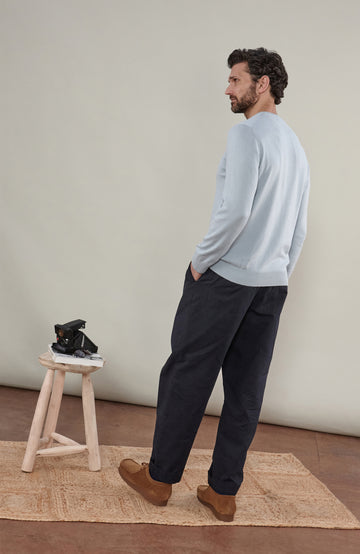 A man wearing a light blue cotton crewneck knit sweater by Paul James Knitwear and dark trousers, photographed against a plain beige studio background.