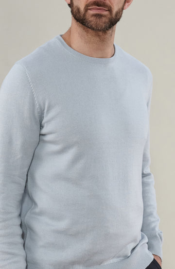 A man wearing a light blue cotton crewneck knit sweater by Paul James Knitwear and dark trousers, photographed against a plain beige studio background.