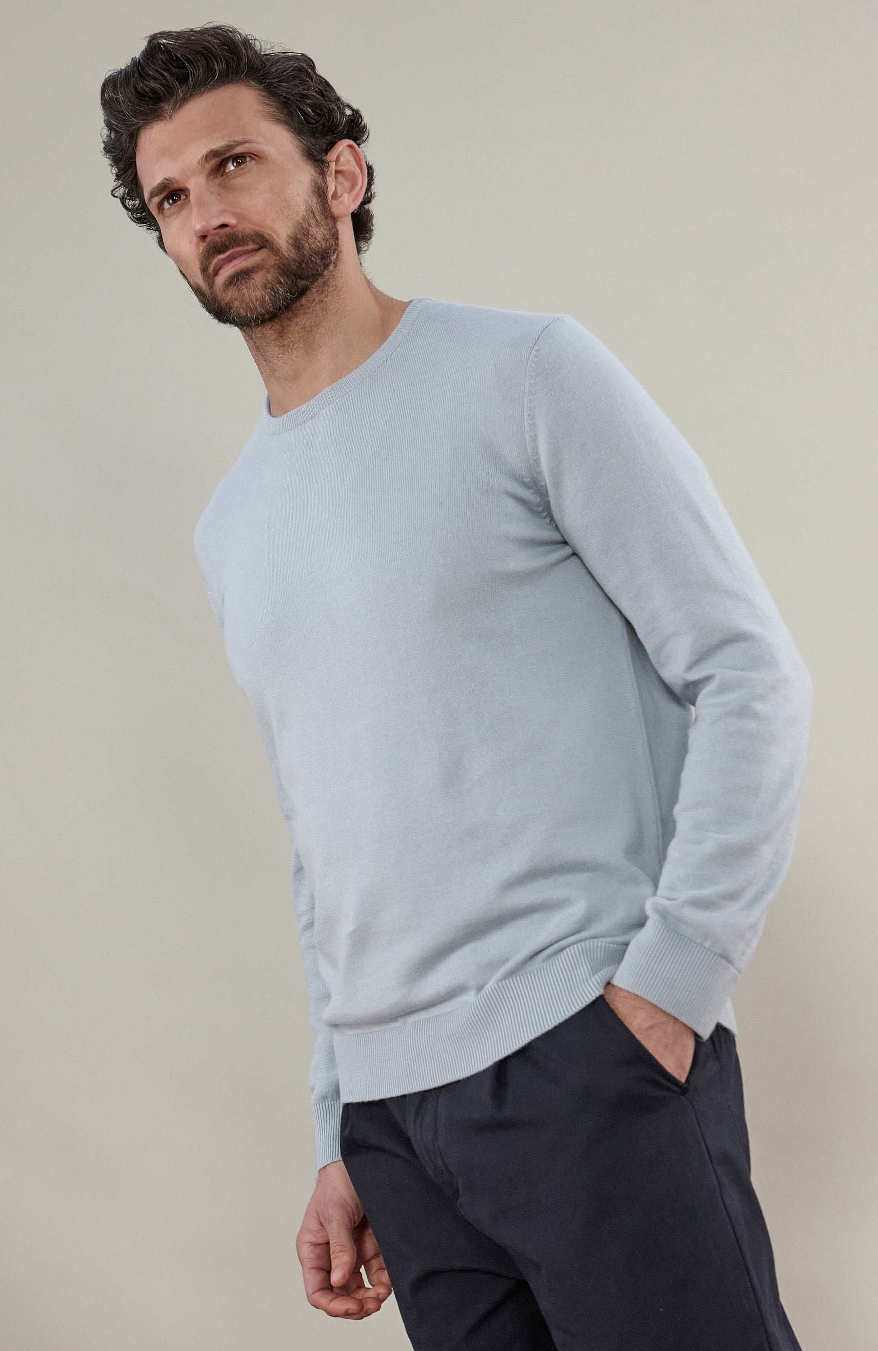 A man wearing a light blue cotton crewneck knit sweater by Paul James Knitwear and dark trousers, photographed against a plain beige studio background.