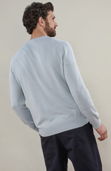 A man wearing a light blue cotton crewneck knit sweater by Paul James Knitwear and dark trousers, photographed against a plain beige studio background.