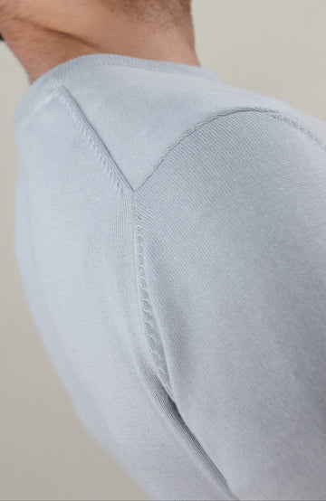 A man wearing a light blue cotton crewneck knit sweater by Paul James Knitwear and dark trousers, photographed against a plain beige studio background.