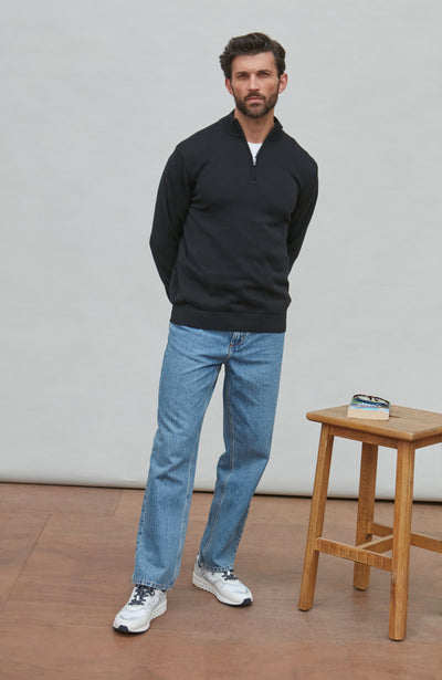 Man wearing a black zip sweater and blue jeans standing in a room with a wooden stool and book.