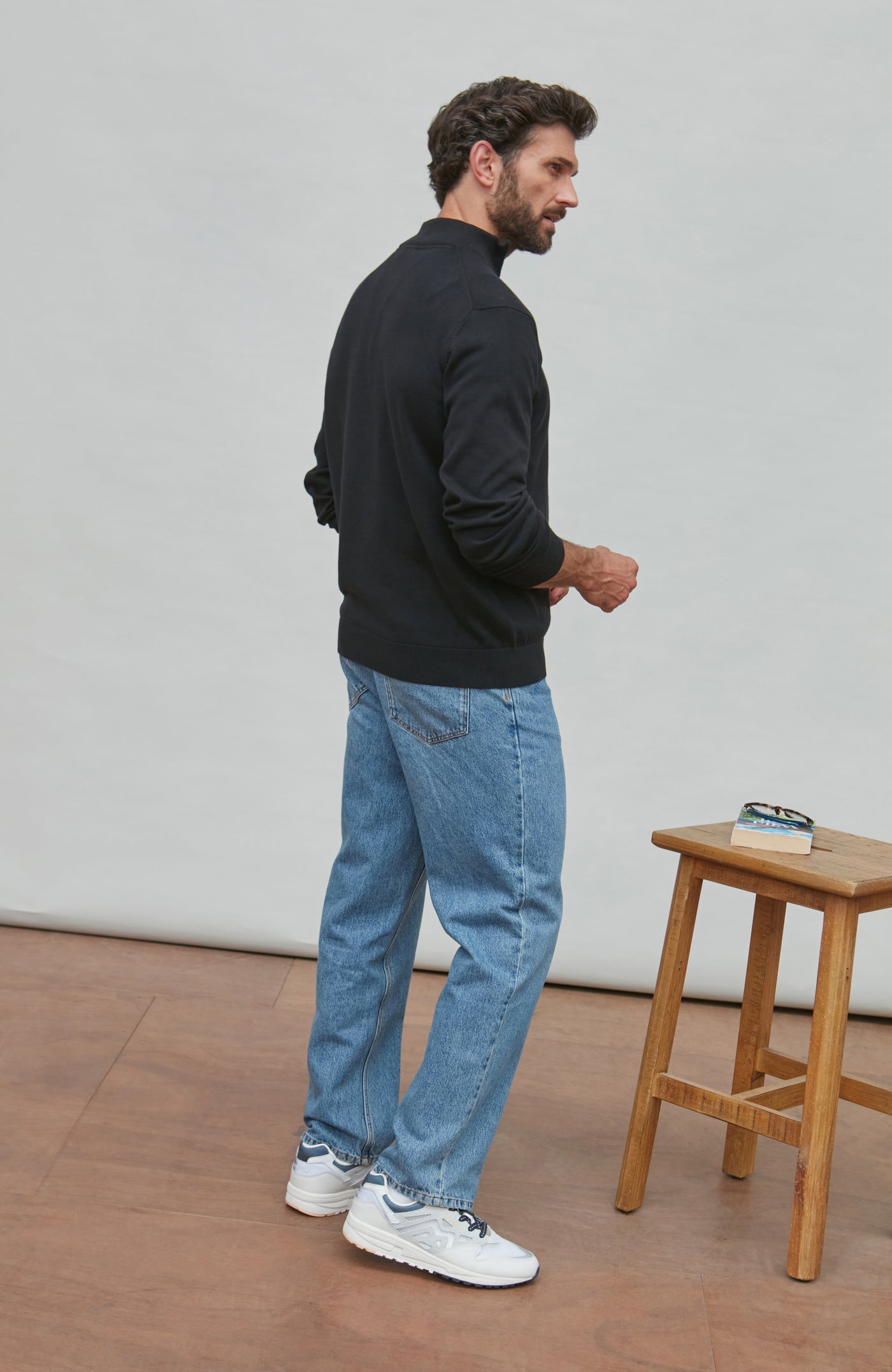 Man wearing a black zip sweater and blue jeans standing in a minimalistic room with a wooden stool.
