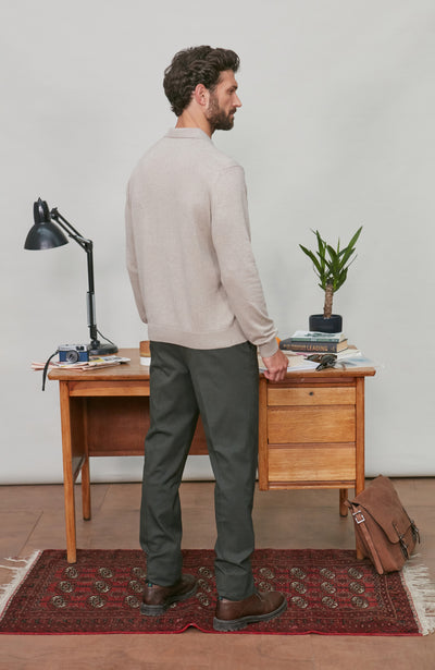 Man wearing polo neck standing in a room with a wooden desk, lamp, and plant.