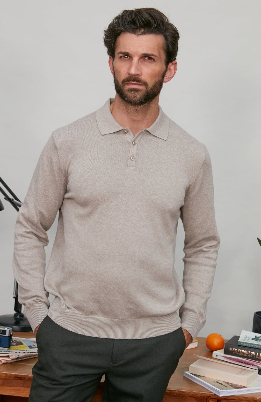 Man wearing a beige polo neck sweater standing in a room with a desk and books.