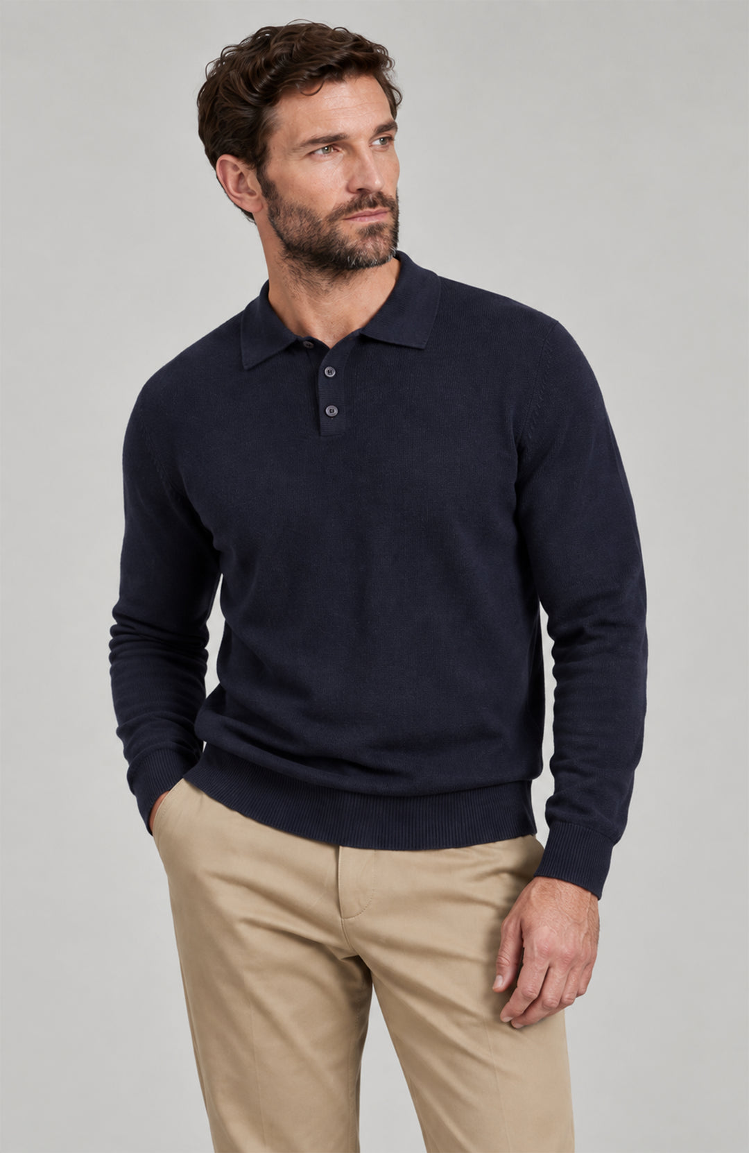 A man wearing a dark navy blue long-sleeve polo sweater and beige trousers, standing against a solid light gray studio background. Made by Paul James Knitwear.
