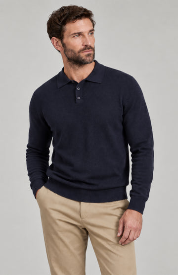 A man wearing a dark navy blue long-sleeve polo sweater and beige trousers, standing against a solid light gray studio background. Made by Paul James Knitwear.