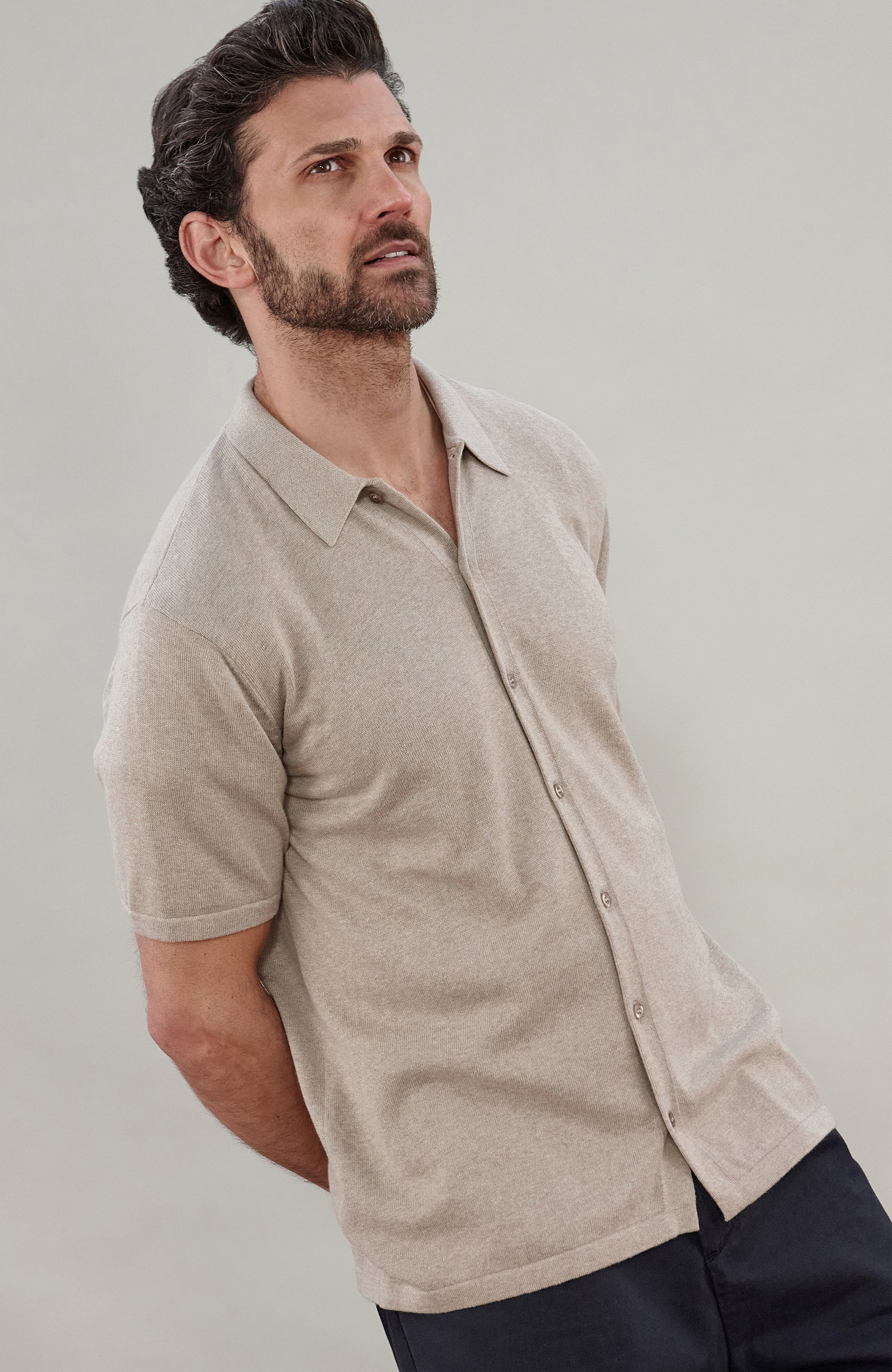 A man wearing a light beige short-sleeve cotton knit button-up shirt by paul james knitwear, shown from the chest down against a plain light gray studio background.