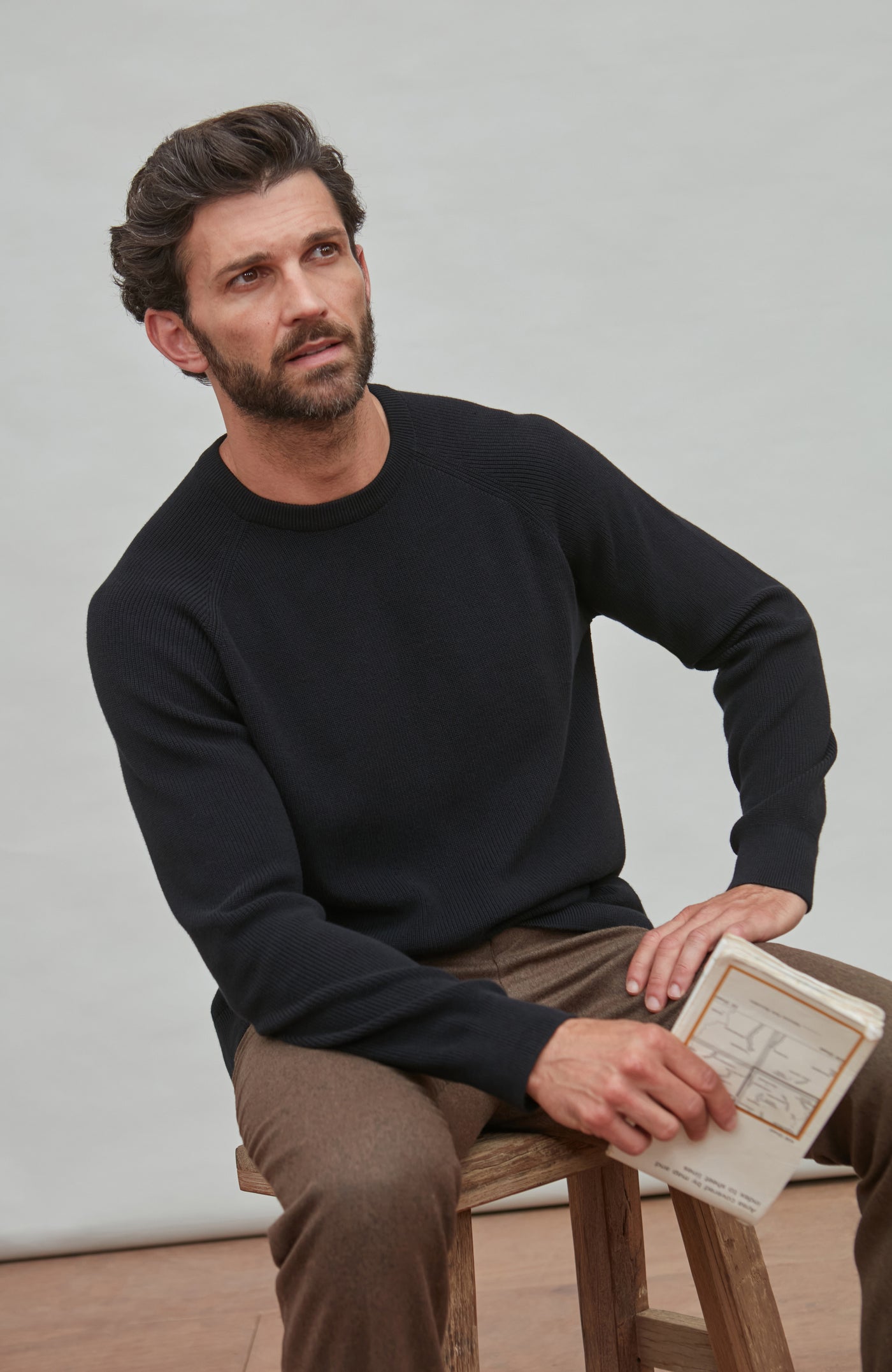 Man wearing black fisherman jumper sitting on a stool holding a book against a plain background