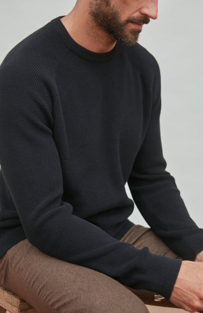 Man wearing a black cotton sweater sitting on a wooden bench against a light gray background
