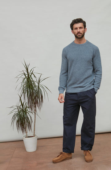 Man wearing a blue cotton rib sweater and navy pants standing next to a potted plant indoors.