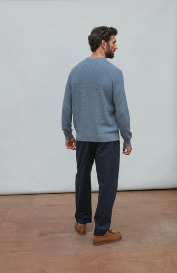 Man wearing blue cotton rib jumper and dark pants standing on a wooden floor with a white wall background