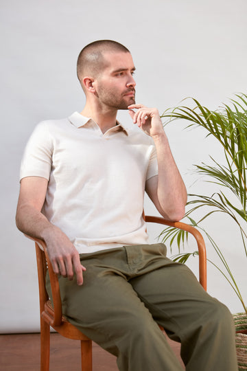 A man sitting in a wooden armchair wearing a short-sleeve white polo shirt by Paul James Knitwear and olive green trousers, with a potted plant in the background against a light gray wall.