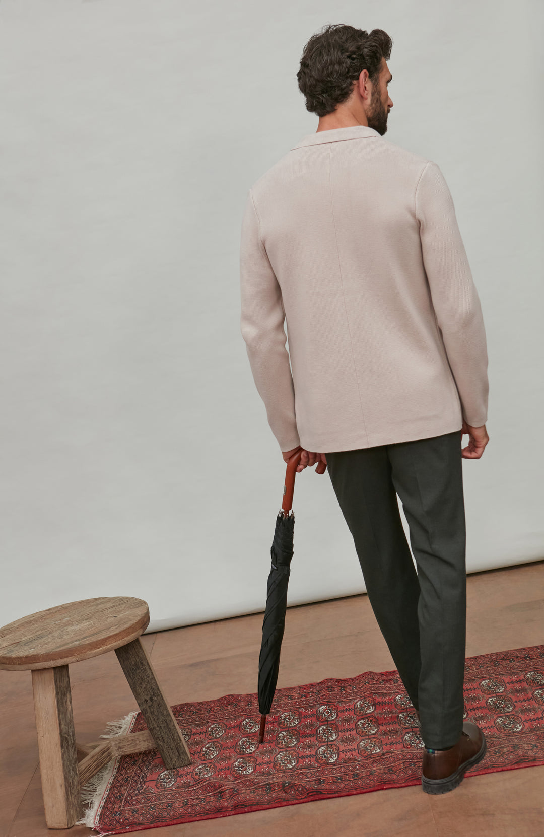 Man holding an umbrella, standing on a patterned rug with a wooden stool nearby.