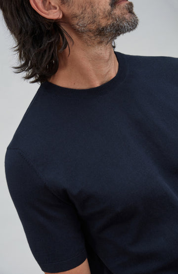 A close-up side view of a man wearing a dark navy blue short-sleeve knit crewneck t-shirt against a light gray background. Made by Paul James Knitwear.