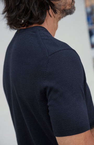 Close-up side view of a man wearing a short-sleeve cotton knit shirt in dark navy blue, showing the shoulder seam and ribbed collar and cuffs. Made by Paul James Knitwear.