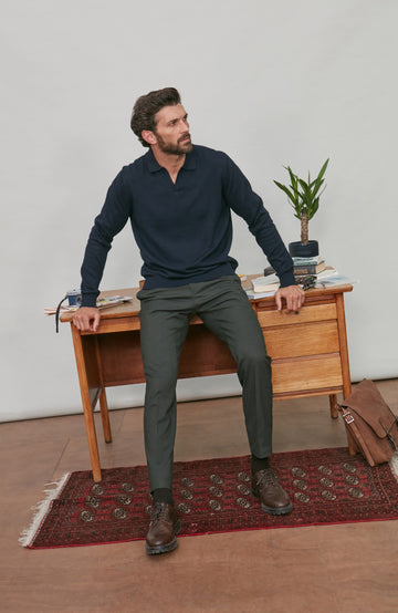 Man leaning against in a navy cotton polo wooden desk in a room with a plant and rug.