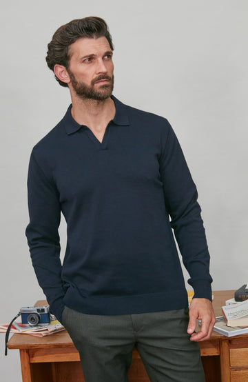 Man wearing a navy long-sleeve cotton polo shirt standing in front of a desk with a camera and books.