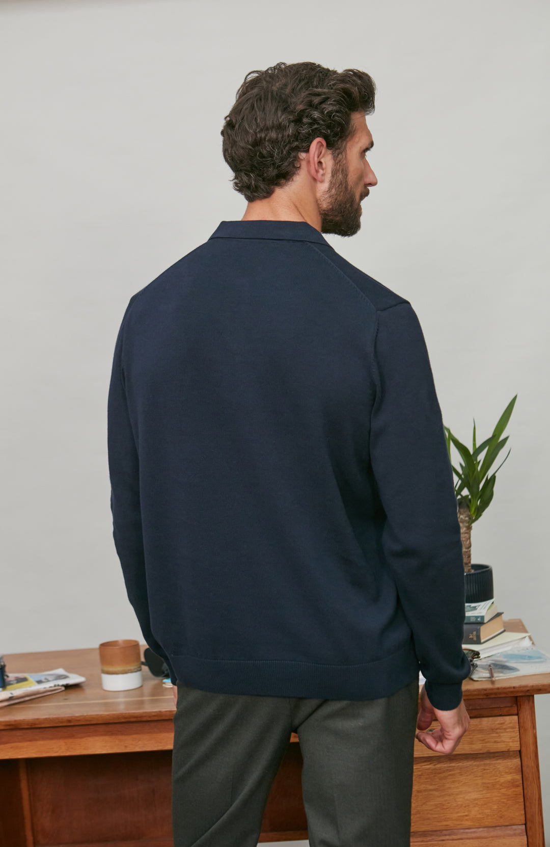 Man wearing a navy cotton polo blue jacket standing in front of a wooden desk with books and a plant.