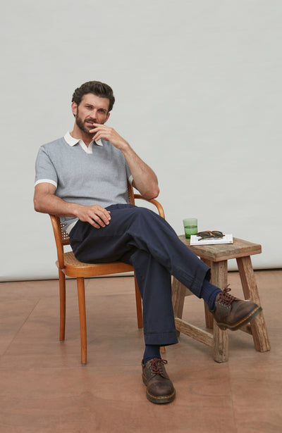 Man wearing nordic summer polo shirt sitting on a wooden chair in a minimalistic room with a wooden stool and small table.