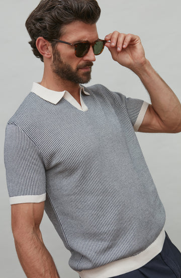Man wearing a patterned polo shirt with a white collar and sunglasses, against a plain background
