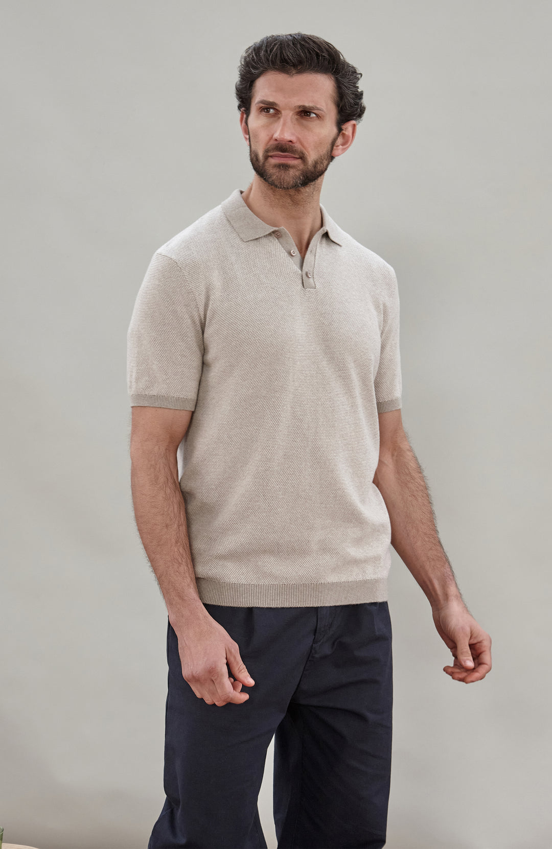 A man wearing a light beige short-sleeve knit polo shirt by Paul James Knitwear  and dark navy trousers, standing against a plain light gray studio background.