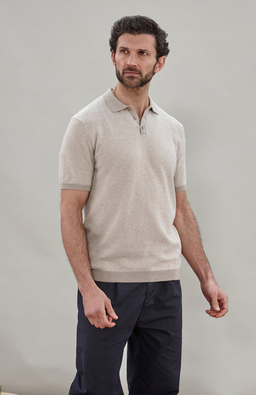 A man wearing a light beige short-sleeve knit polo shirt by Paul James Knitwear  and dark navy trousers, standing against a plain light gray studio background.