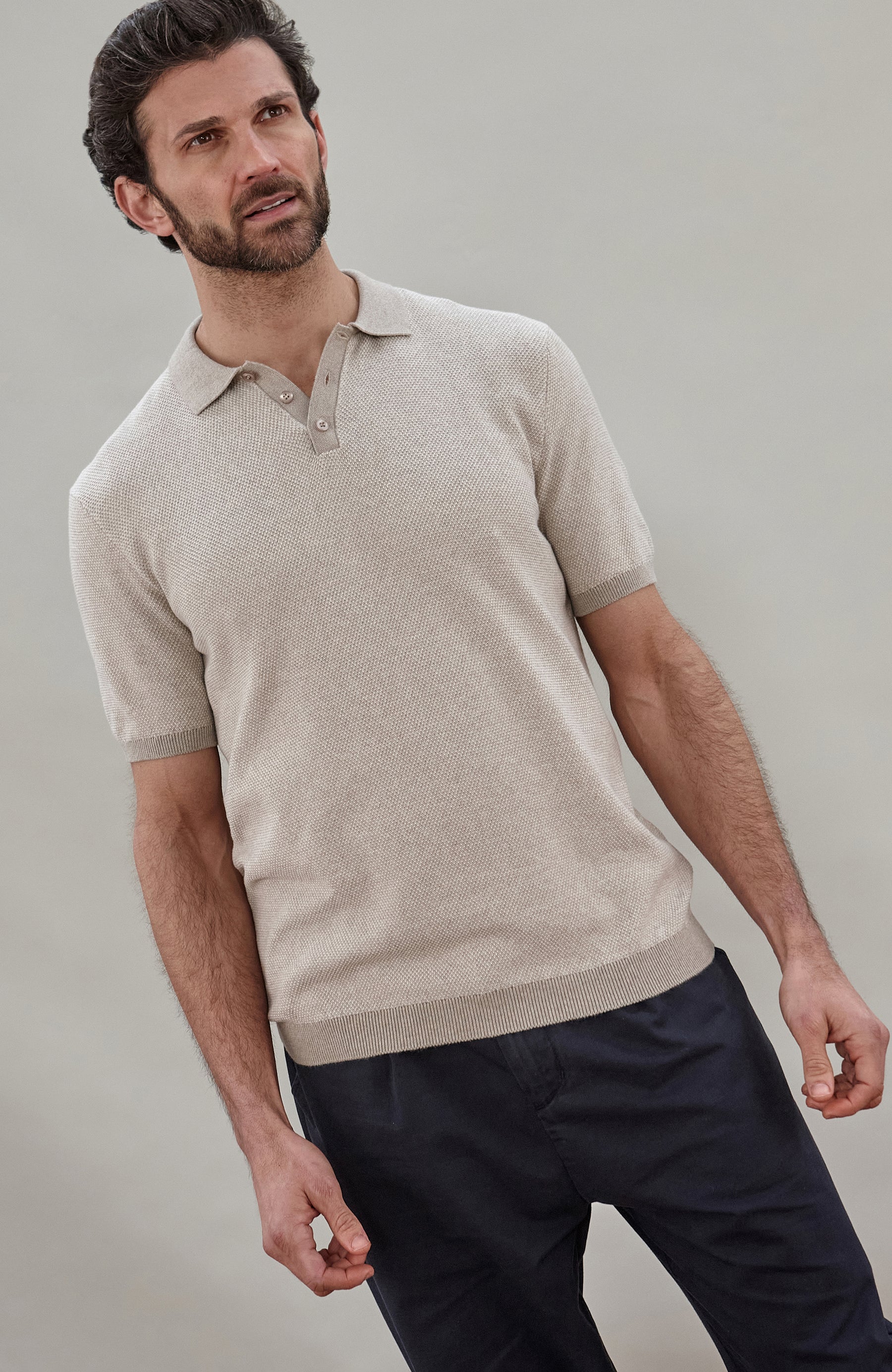 A man wearing a light beige short-sleeve knit polo shirt by Paul James Knitwear and dark navy trousers, photographed against a plain light gray studio background.