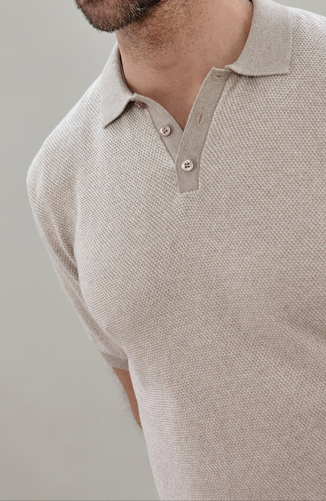 Close-up of a man wearing a light beige textured short-sleeve polo shirt by Paul James Knitwear with a two-button placket.