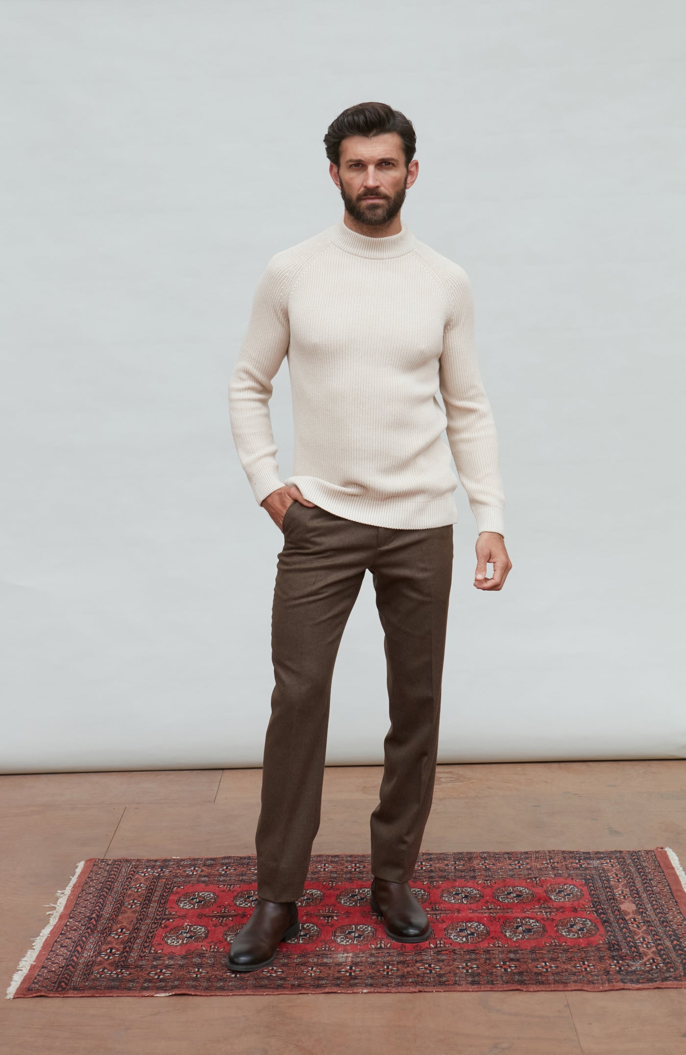 Man wearing a beige sweater and brown pants standing on a red patterned rug with a white wall background