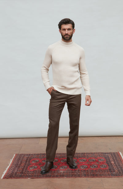 Man wearing a beige sweater and brown pants standing on a red patterned rug with a white wall background