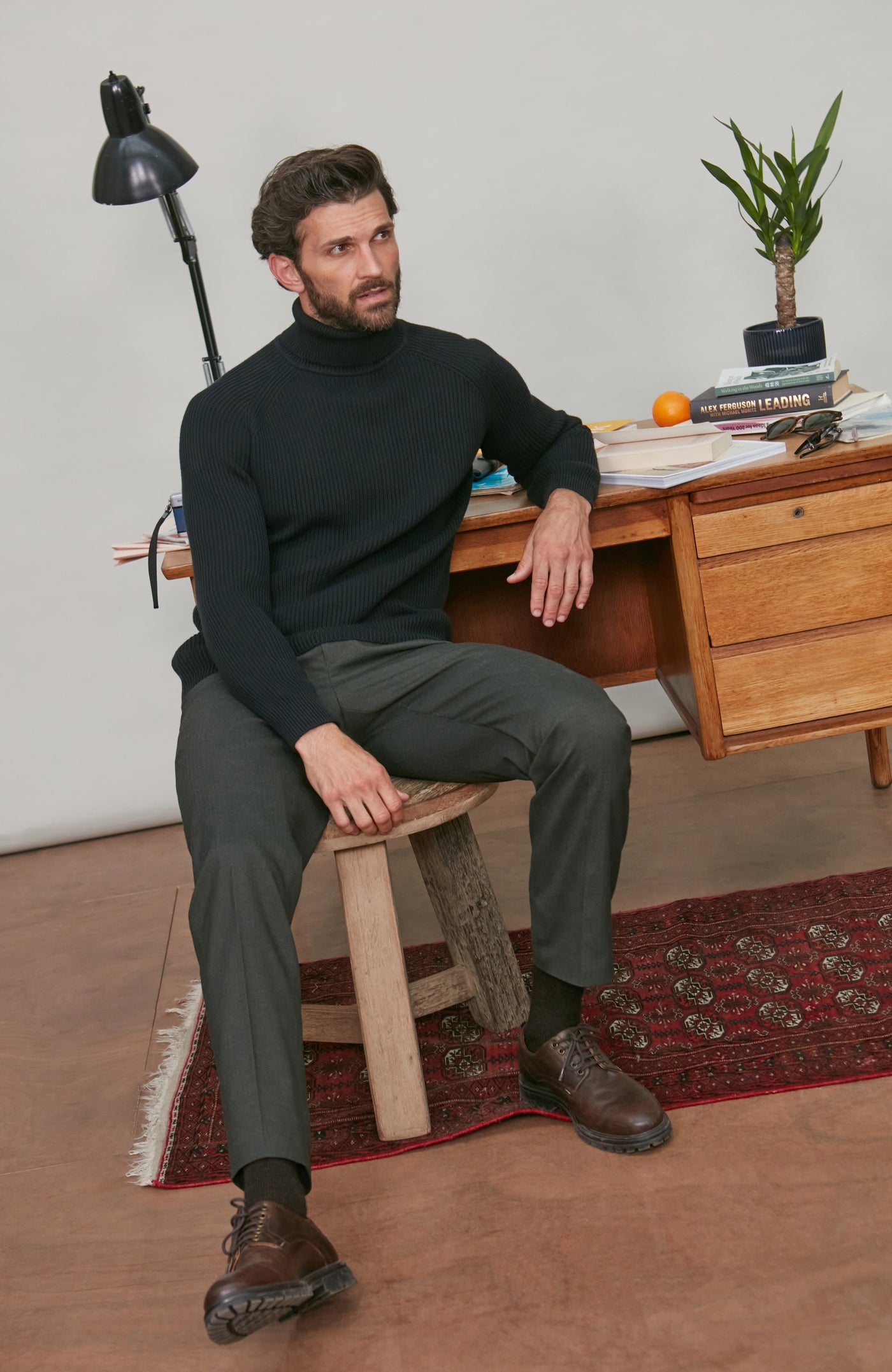Man wearing black ribbed turtleneck sweater on a wooden stool in front of a desk with a plant and lamp.