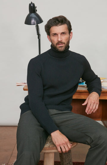 Man wearing a black ribbed turtleneck sweater and gray pants sitting on a wooden stool.