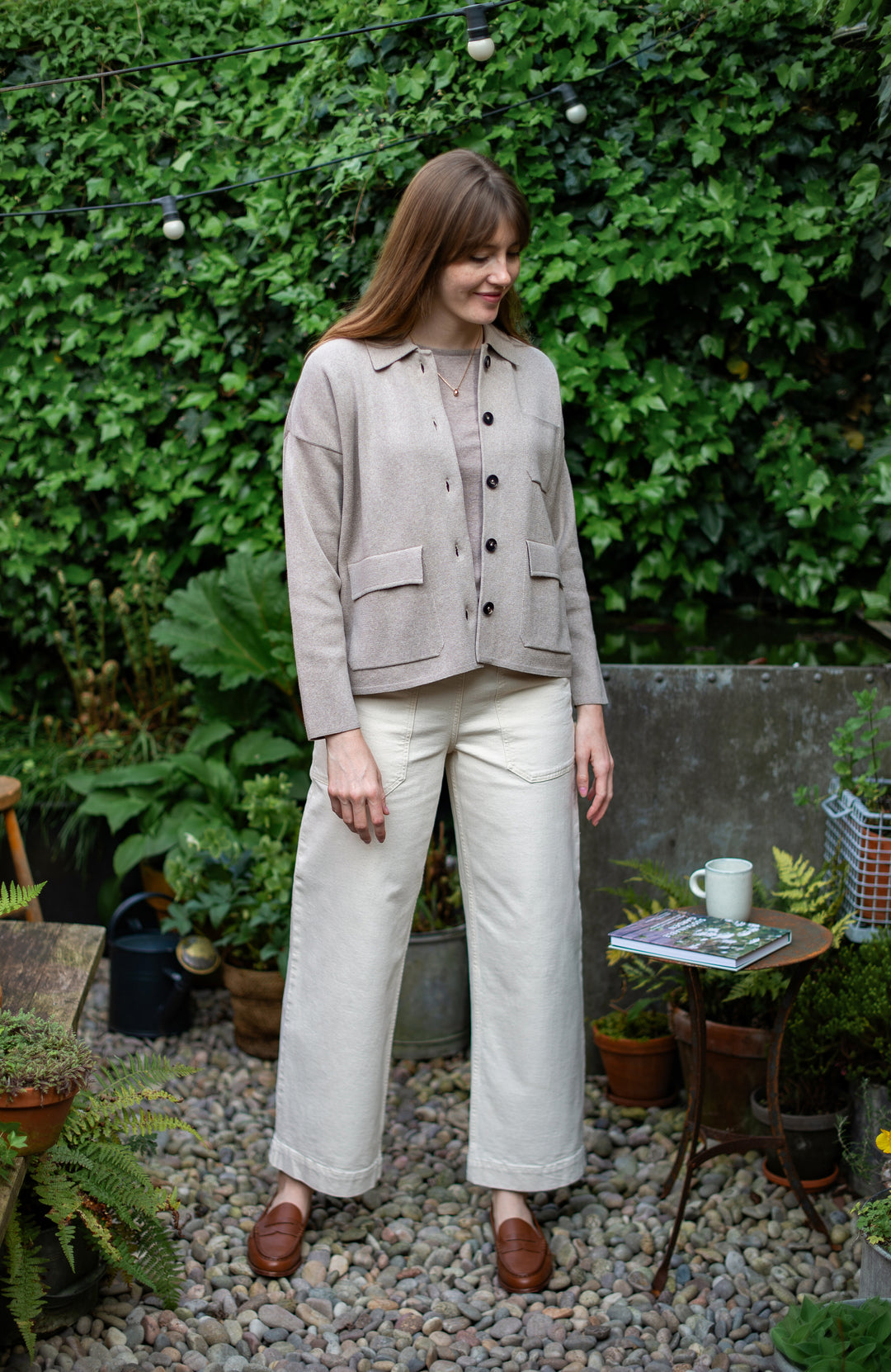 Woman standing in a garden wearing a beige jacket and white pants.