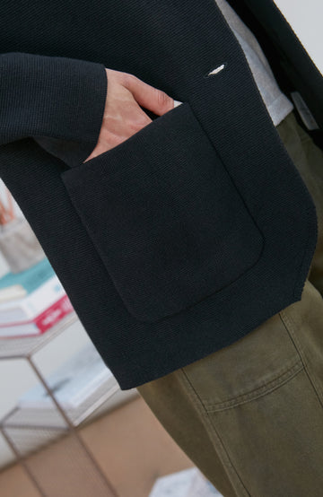 A model wearing a black knitted blazer by Paul James Knitwear over a light gray crewneck t-shirt and olive green pants, standing in a minimalist room with a white wall and a small wire shelf.