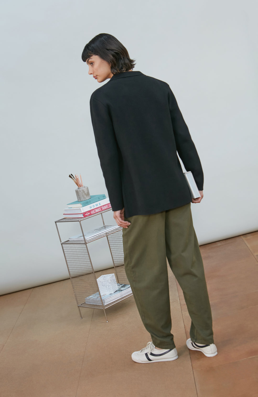 A model wearing a black knitted blazer by Paul James Knitwear over a light gray crewneck t-shirt and olive green pants, standing in a minimalist room with a white wall and a small wire shelf.