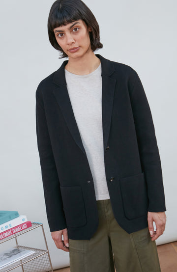 A model wearing a black knitted blazer by Paul James Knitwear over a light gray crewneck t-shirt and olive green pants, standing in a minimalist room with a white wall and a small wire shelf.