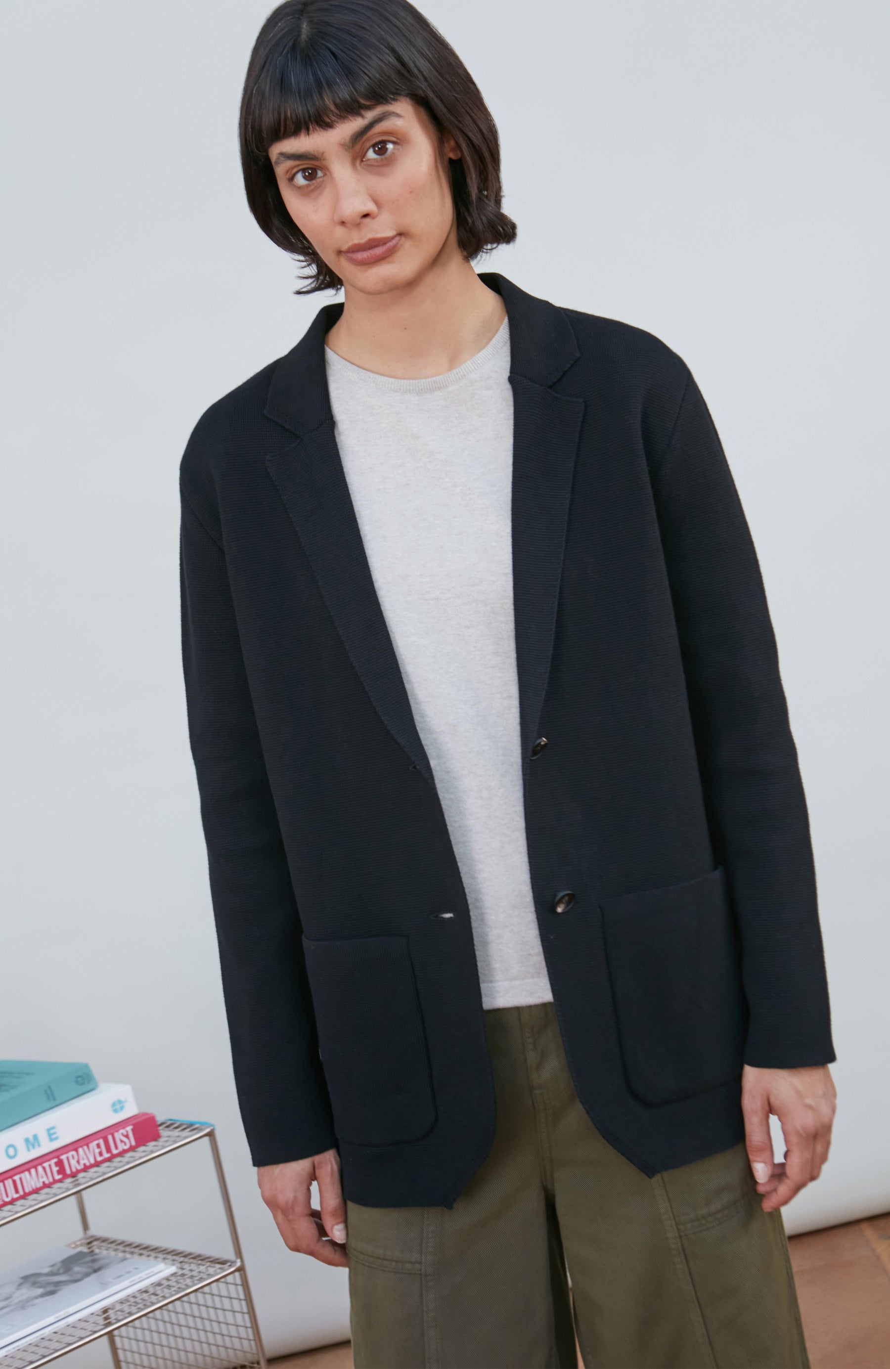 A model wearing a black knitted blazer by Paul James Knitwear over a light gray crewneck t-shirt and olive green pants, standing in a minimalist room with a white wall and a small wire shelf.