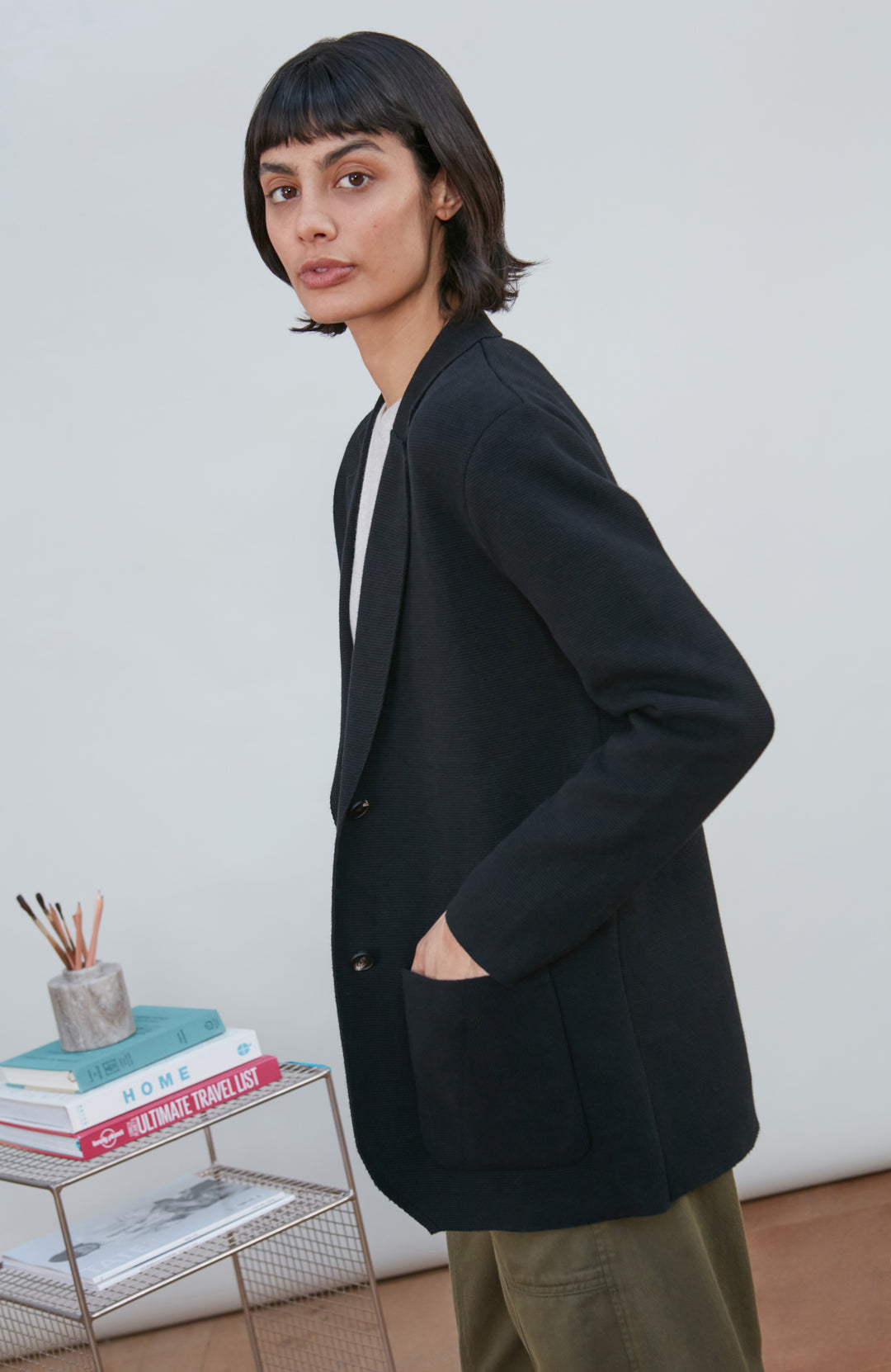 A model wearing a black knitted blazer by Paul James Knitwear over a light gray crewneck t-shirt and olive green pants, standing in a minimalist room with a white wall and a small wire shelf.