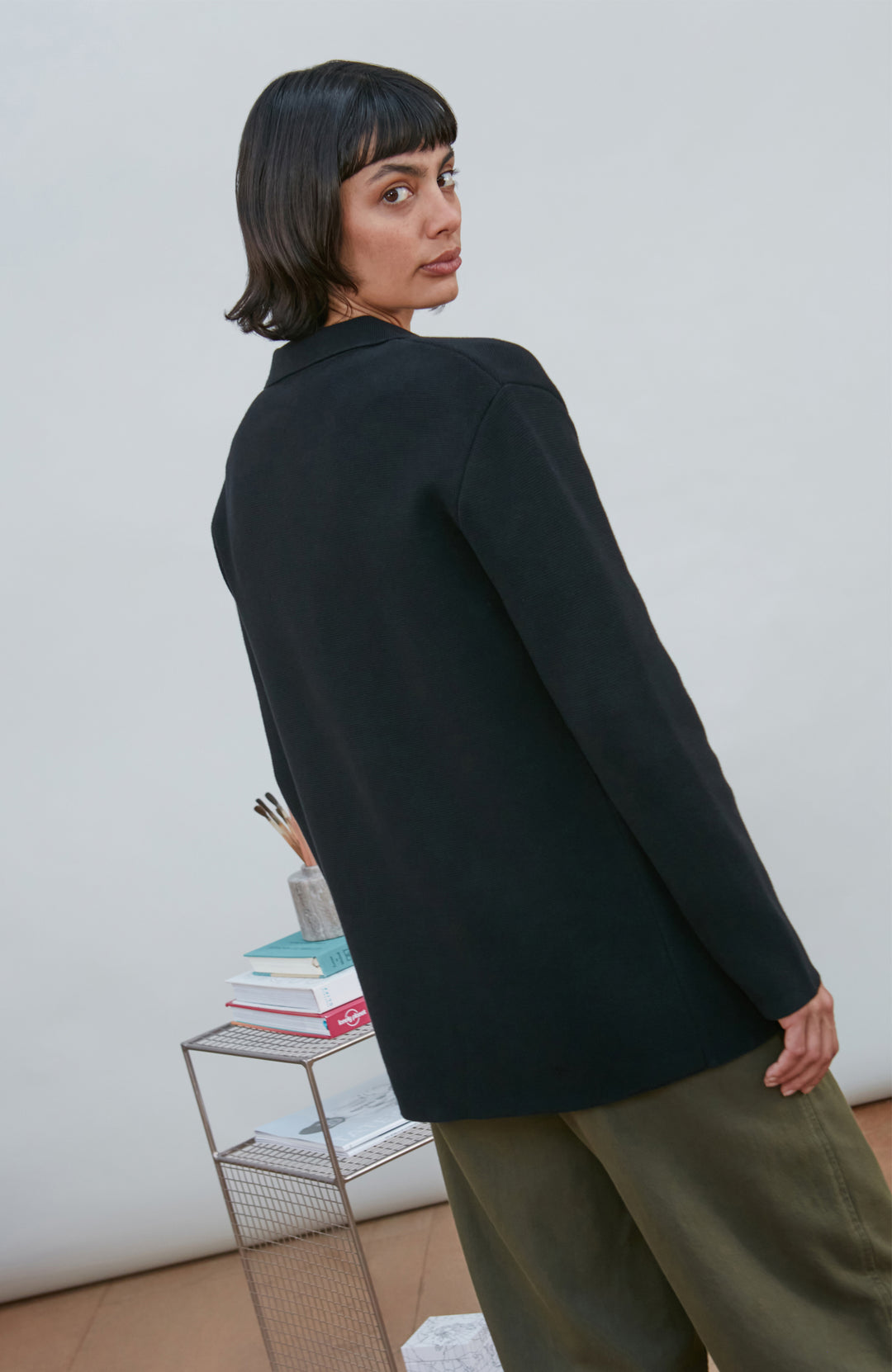 A model wearing a black knitted blazer by Paul James Knitwear over a light gray crewneck t-shirt and olive green pants, standing in a minimalist room with a white wall and a small wire shelf.
